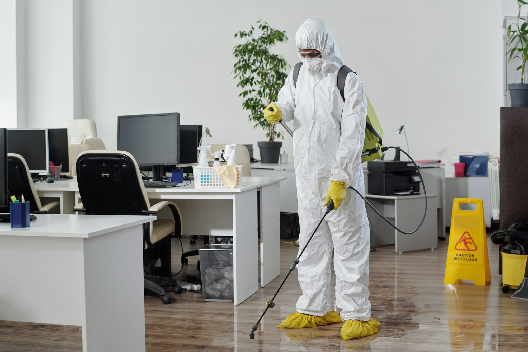 Male worker in a hazmat suit disinfecting an office floor with cleaning equipment, emphasizing safety and hygiene.